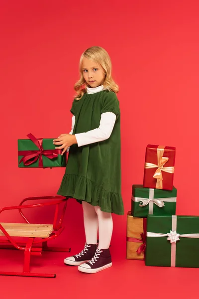 Girl in green dress holding present near sled and stack of gift boxes on red — Stock Photo