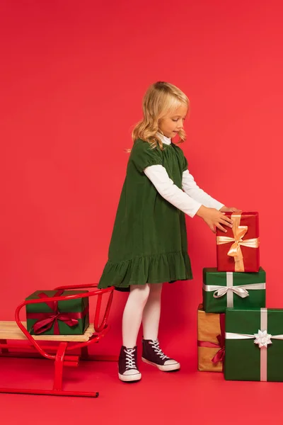 Child in green dress putting gift on stack of gift boxes near sled on red — Stock Photo