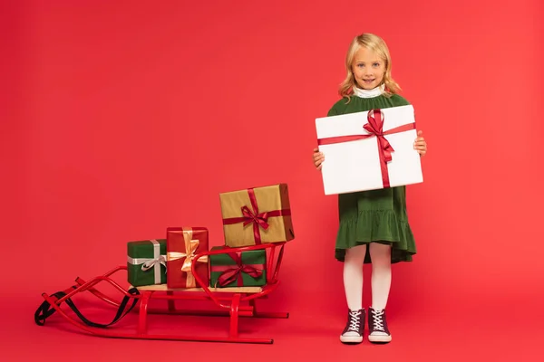 Full length view of stylish girl with present near sled with gift boxes on red — Stock Photo