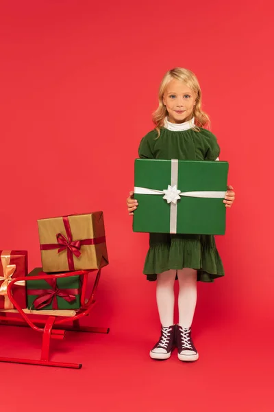Positive girl holding green gift box near sled with presents on red — Stock Photo