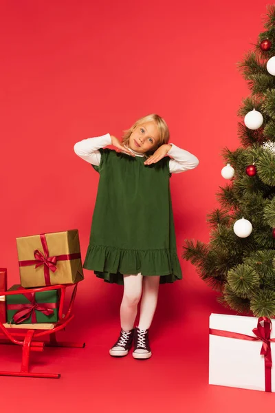 Ganzkörperansicht von Kind in grünem Kleid posiert in der Nähe von Weihnachtsbaum und Schlitten mit Geschenkschachteln auf rot — Stockfoto