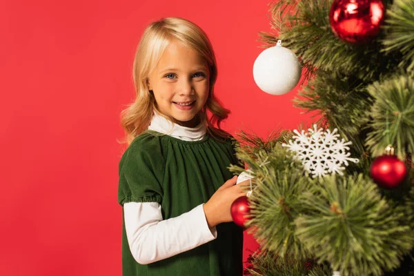 Positive girl in green dress smiling at camera near decorated christmas tree isolated on red — Stock Photo