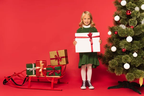 Niña feliz sosteniendo presente cerca del árbol de Navidad y trineo con cajas de regalo en rojo - foto de stock