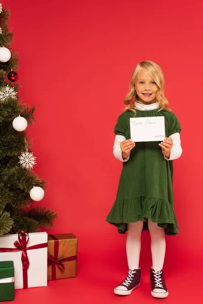 Smiling girl with letter to santa clause near presents under christmas tree on red — Stock Photo