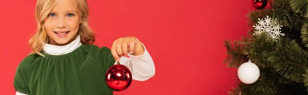 Lächelndes Kind mit glänzender Weihnachtskugel neben geschmückter Fichte isoliert auf rotem Grund, Banner — Stockfoto