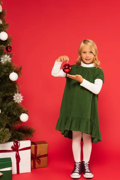 Щаслива дитина тримає різдвяний м'яч біля прикрашеної ялинки та подарункових коробок на червоному — стокове фото