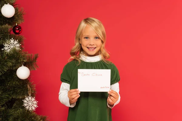 Alegre chica celebración carta a santa cláusula cerca decorado árbol de Navidad aislado en rojo - foto de stock