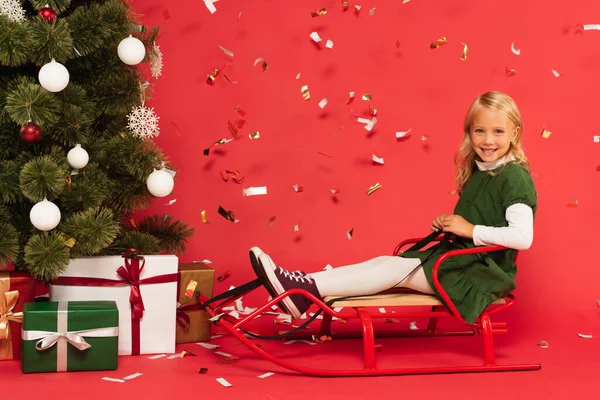 Cheerful girl in green dress sitting on sled near gift boxes under christmas tree on red — Stock Photo