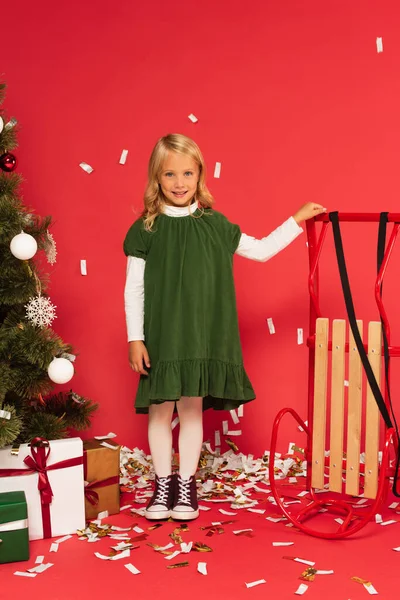 Gaie fille en robe verte près traîneau et cadeaux sous arbre de Noël sur rouge — Photo de stock