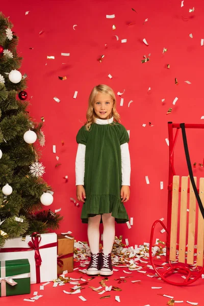 Glückliches Mädchen in grünem Kleid und Turnschuhen lächelt in die Kamera neben Schlitten und Geschenke unter dem Weihnachtsbaum auf rot — Stockfoto