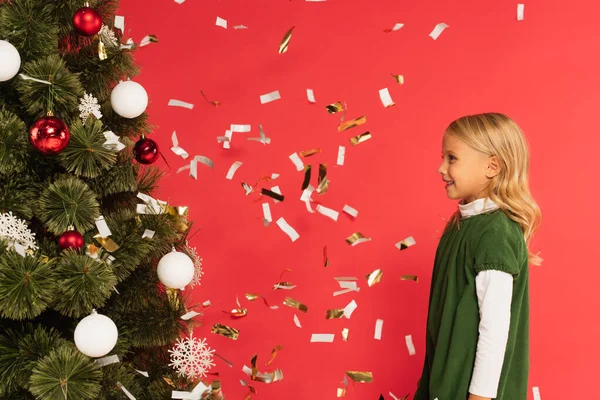 Vista lateral da menina feliz em vestido verde perto da árvore de Natal sob queda confete isolado no vermelho — Fotografia de Stock