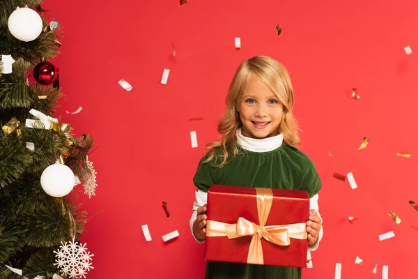 Happy kid with gift box smiling at camera near christmas tree under falling confetti isolated on red — Stock Photo