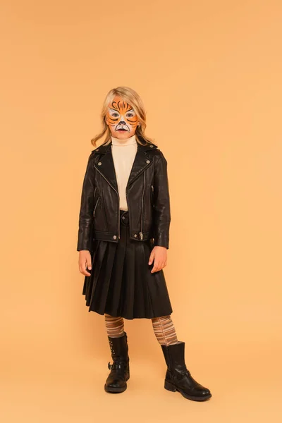 Menina na pintura de rosto de tigre, jaqueta de couro preto, botas e saia plissada posando no bege — Fotografia de Stock