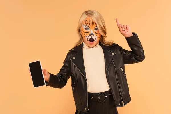 Chica sorprendida en el maquillaje tigre mostrando teléfono inteligente con pantalla en blanco y apuntando con el dedo aislado en beige - foto de stock