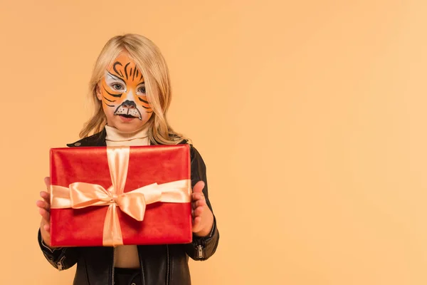 Fille en tigre maquillage tenant boîte cadeau tout en regardant la caméra isolée sur beige — Photo de stock