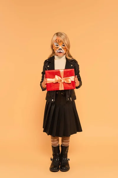 Повнометражний вигляд дитини в стильному одязі і тигровому макіяжі, що тримає подарунок на бежевому — стокове фото