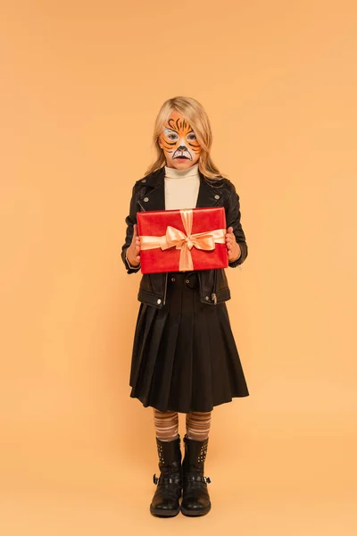 Vue pleine longueur de l'enfant dans des vêtements élégants et le maquillage du tigre tenant présent sur beige — Photo de stock