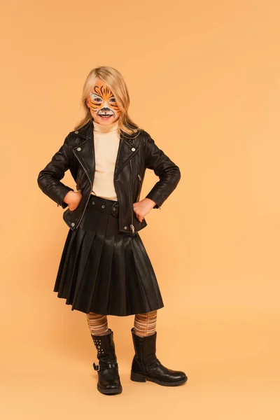 Alegre chica en tigre maquillaje, chaqueta de cuero y botas posando con las manos en las caderas en beige - foto de stock