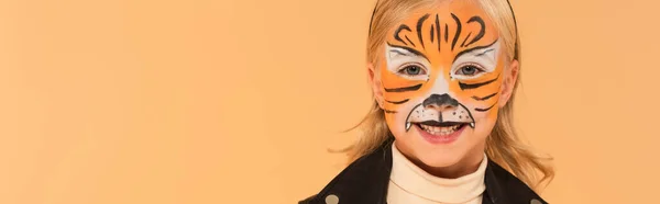 Радісна дитина з тигровим малюнком обличчя, посміхається на камеру ізольовано на бежевому, банер — стокове фото