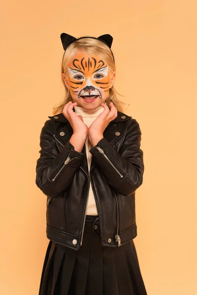 Lächelndes Kind in schwarzer Lederjacke und Tiger-Make-up, das Hände in Gesichtsnähe hält, isoliert auf beige — Stockfoto