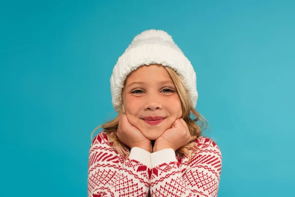 Glückliches Kind in Strickmütze und warmem Pullover mit Ornament, das isoliert auf blauem Grund in die Kamera blickt — Stockfoto