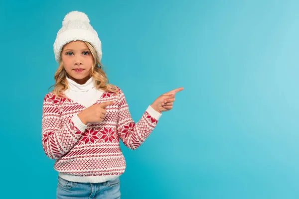 Girl in knitted hat and sweater looking at camera while pointing aside with fingers isolated on blue — Stock Photo