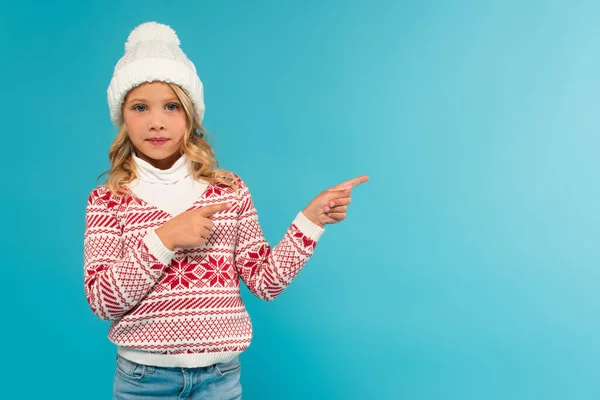 Fille en bonnet tricoté et pull regardant caméra tout en pointant de côté avec les doigts isolés sur bleu — Photo de stock