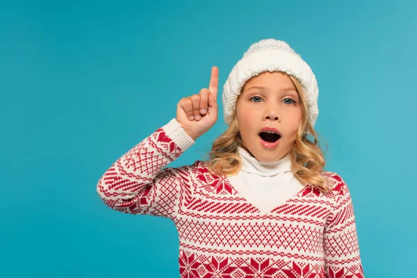 Astonished child in warm sweater and hat looking at camera and pointing up with finger isolated on blue — Stock Photo