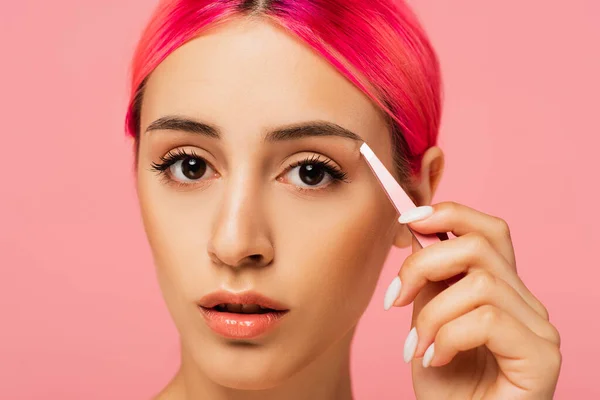 Jeune femme aux cheveux colorés tenant des pinces à épiler tout en façonnant le sourcil isolé sur rose — Photo de stock