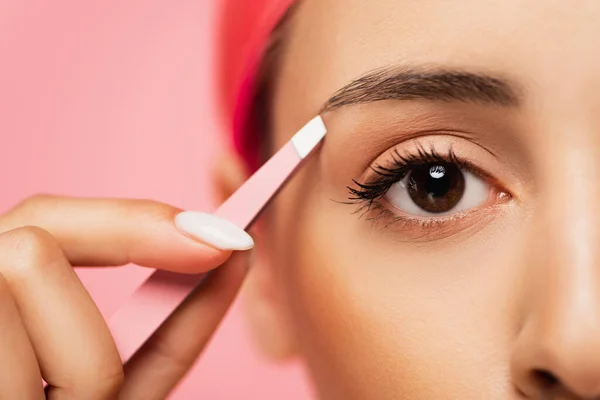 Vue recadrée d'une jeune femme aux cheveux teints tenant une pince à épiler tout en formant un sourcil isolé sur rose — Photo de stock