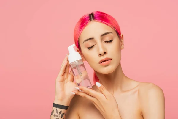 Jeune femme tatouée avec des cheveux colorés et les yeux fermés tenant bouteille avec un produit nettoyant isolé sur rose — Photo de stock