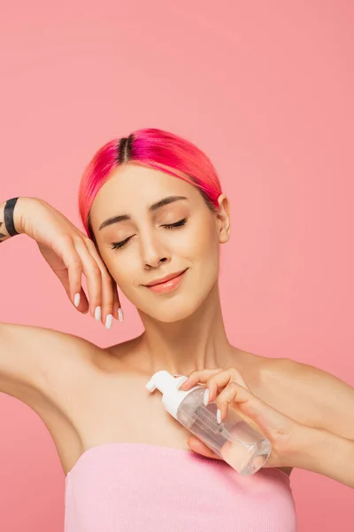 Giovane donna tatuata con capelli colorati e occhi chiusi che tiene in bottiglia con prodotto detergente mentre sorride isolata sul rosa — Foto stock