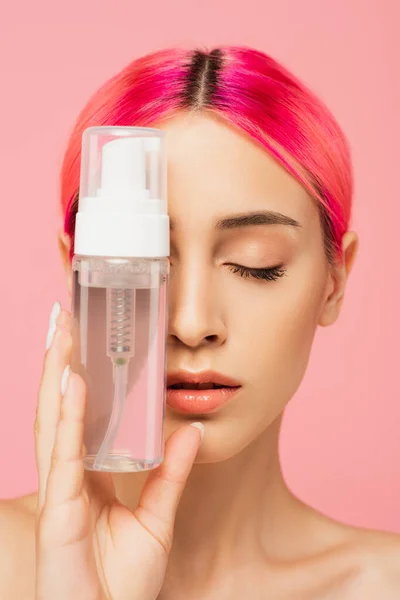 Mujer joven con el pelo colorido y los ojos cerrados sosteniendo botella con producto de limpieza aislado en rosa - foto de stock