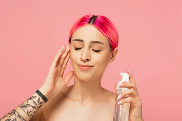 Mujer joven tatuada con el pelo teñido y los ojos cerrados sosteniendo botella con producto de limpieza mientras sonríe aislado en rosa - foto de stock