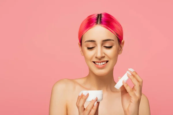 Cheerful young woman with dyed hair looking at container with cosmetic cream isolated on pink — Stock Photo