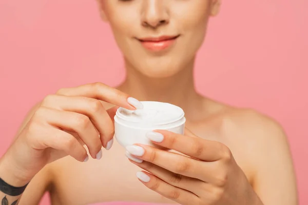 Vista recortada de mujer joven tatuada sonriendo mientras sostiene contenedor con crema cosmética aislada en rosa - foto de stock