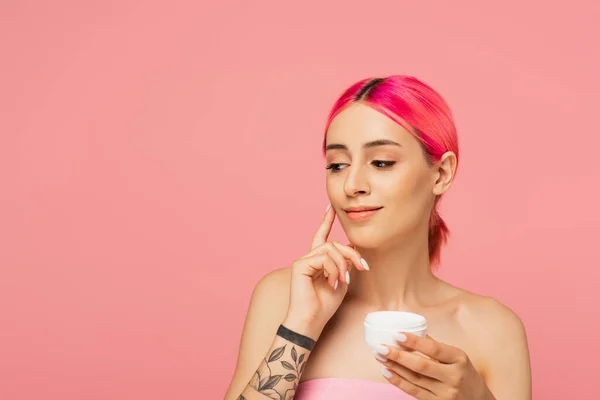 Mujer joven tatuada con pelo colorido sonriendo mientras sostiene el recipiente y aplica crema facial aislada en rosa - foto de stock