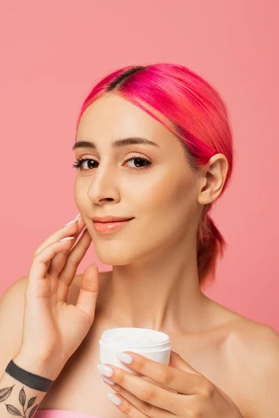 Jovem tatuada com cabelo tingido sorrindo enquanto segurando recipiente e aplicando creme facial isolado em rosa — Fotografia de Stock