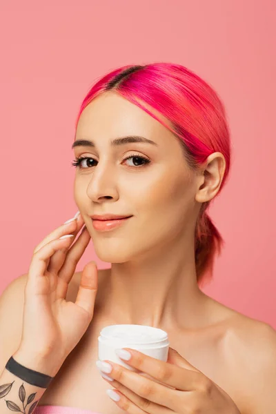 Mujer joven tatuada con el pelo teñido sonriendo mientras sostiene el recipiente y la aplicación de crema facial aislado en rosa - foto de stock