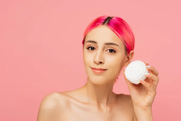 Mujer joven positiva con el pelo colorido sonriendo mientras sostiene el envase con crema facial aislado en rosa - foto de stock