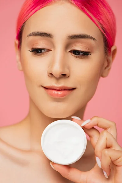 Jovem mulher positiva com cabelo tingido segurando recipiente com creme cosmético isolado em rosa — Fotografia de Stock