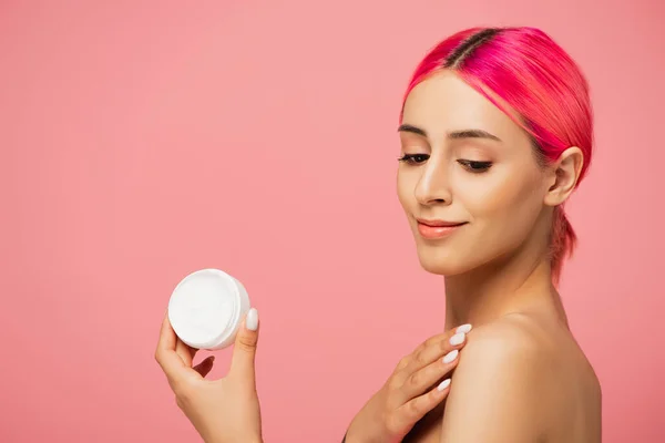 Positive junge Frau mit gefärbten Haaren hält Behälter mit kosmetischer Creme und lächelt isoliert auf rosa — Stockfoto