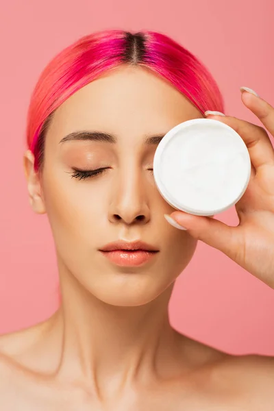 Mujer joven con el ojo de cubierta de pelo colorido mientras sostiene el envase con crema cosmética aislada en rosa - foto de stock