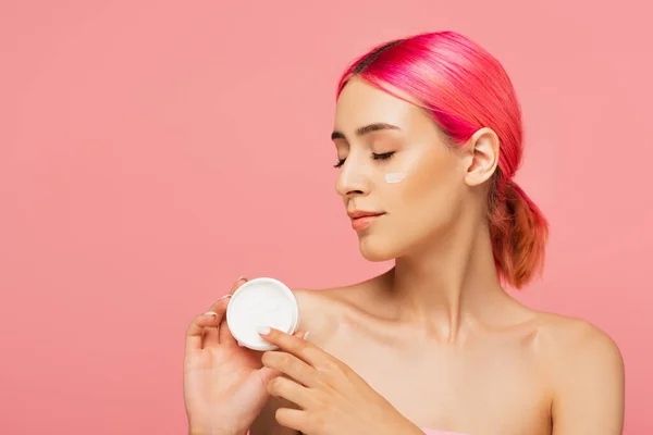 Mujer joven con el pelo teñido y crema facial en la mejilla sosteniendo contenedor aislado en rosa - foto de stock