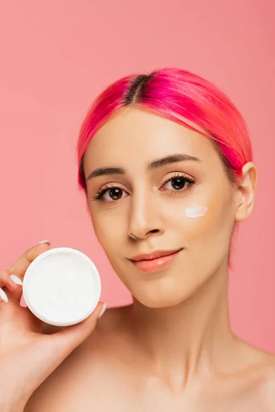 Mujer joven complacida con el pelo teñido y crema facial en la mejilla sosteniendo contenedor aislado en rosa - foto de stock