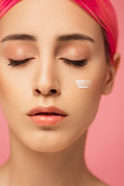 Primer plano de la mujer joven con los ojos cerrados y crema facial en la mejilla aislado en rosa - foto de stock