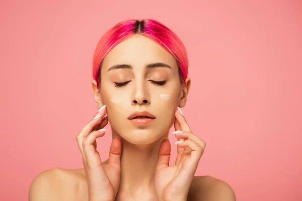 Young woman with closed eyes and face cream touching cheeks isolated on pink — Stock Photo