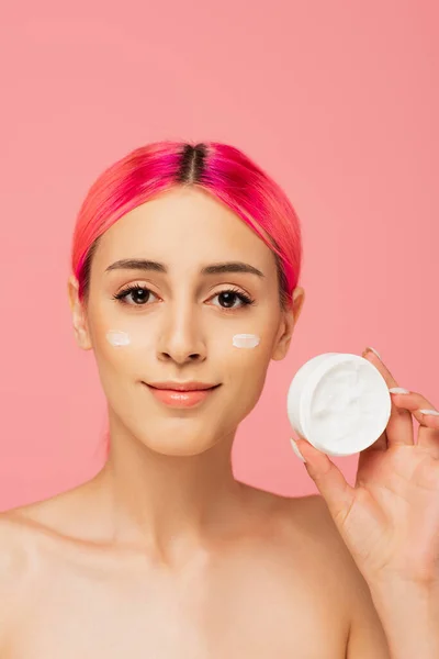 Joyful young woman with colorful hair and cream on cheeks holding container isolated on pink — Stock Photo
