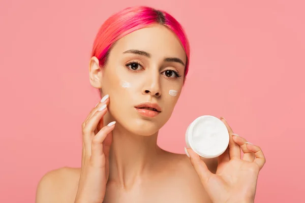 Mujer joven con pelo colorido y crema en las mejillas sosteniendo contenedor aislado en rosa - foto de stock