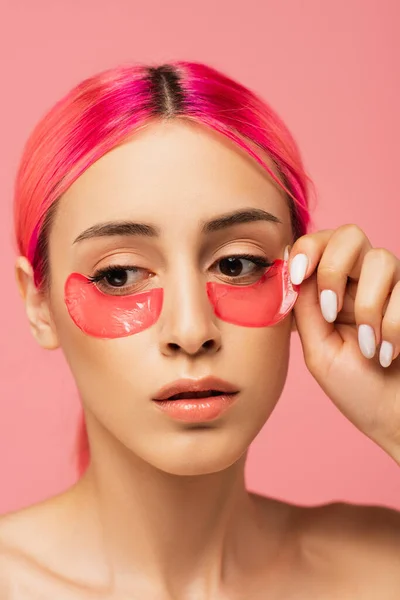 Mujer joven con pelo colorido aplicando parches para los ojos de colágeno aislados en rosa - foto de stock